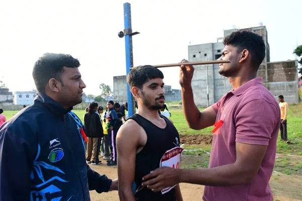 मैदान में एक युवक प्रतिभागी के सिर के पास मापने वाली छड़ लगाए हुए है और दूसरा व्यक्ति पास खड़ा है