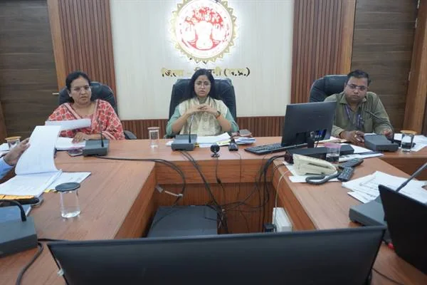 बैठक कक्ष में मेज के सामने बैठीं तीन अधिकारी, सामने कंप्यूटर, माइक्रोफोन और दस्तावेज रखे हैं
