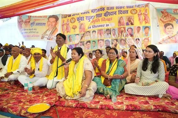पंडाल में दरी पर बैठे मंत्री संपतिया उइके माइक पर बोलती हुईं, साथ में पीले दुपट्टे पहने जनप्रतिनिधि और ग्रामीण; पीछे राजा शंकर शाह और कुंवर रघुनाथ शाह के चित्र वाले बैनर लगे हैं।