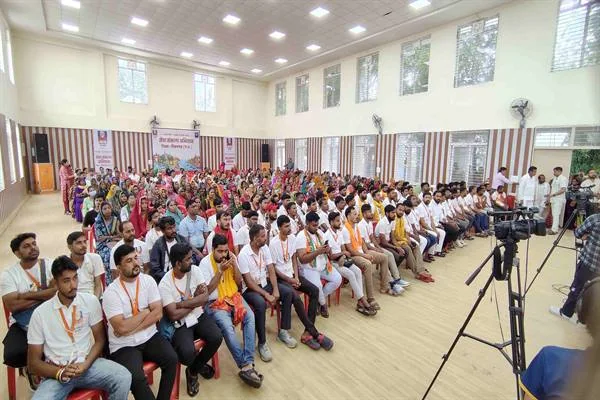 एक बड़े हॉल में कुर्सियों पर बैठे सैकड़ों लोग, आगे की पंक्तियों में सफेद टी-शर्ट और गले में पट्टा पहने युवा, पीछे साड़ी पहने महिलाएं, दाईं ओर तिपाई पर लगा वीडियो कैमरा।